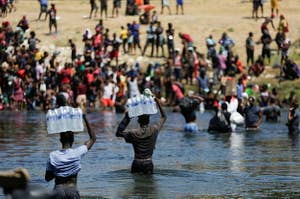 Two people carrying cases of water on their head cross back over the river to a crowd of people waiting