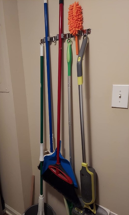 A wall-mounted organizer with 4 hooks and 3 racks holding a broom, mop, Swiffer, and duster