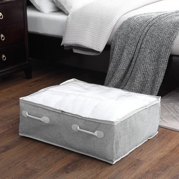Gray storage bin with white handles and top sitting on the floor next to a bed.