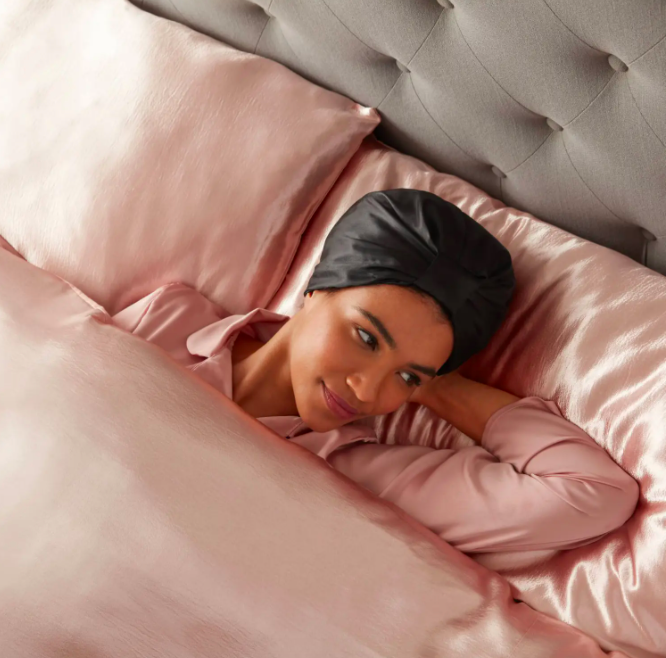 a smiling person laying in bed while wearing the satin bonnet