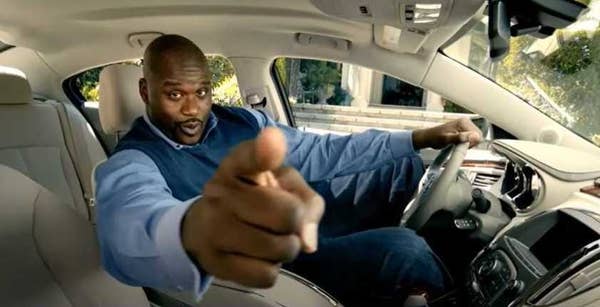 Shaq in a car and points at the camera