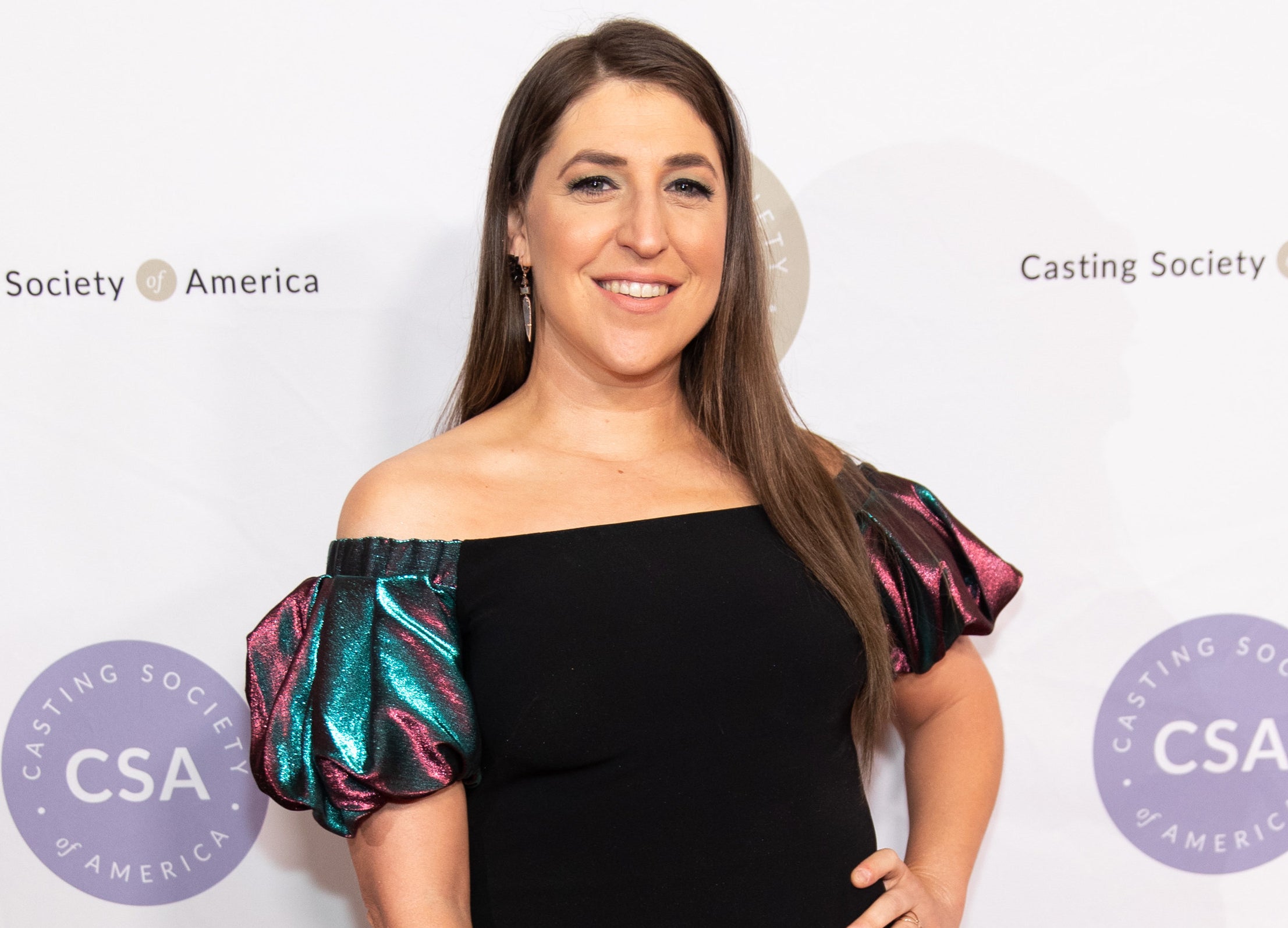 Mayim smiles while wearing an off the shoulder dress at an event