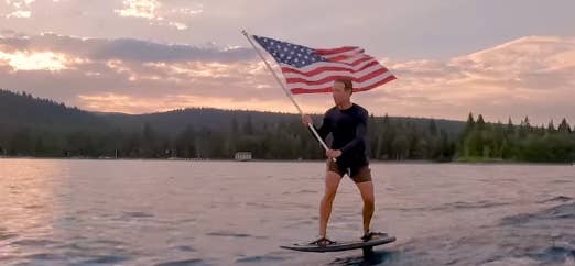 In this still, Mark Zuckerberg is riding a hydrofoil on the water and holding an American flag