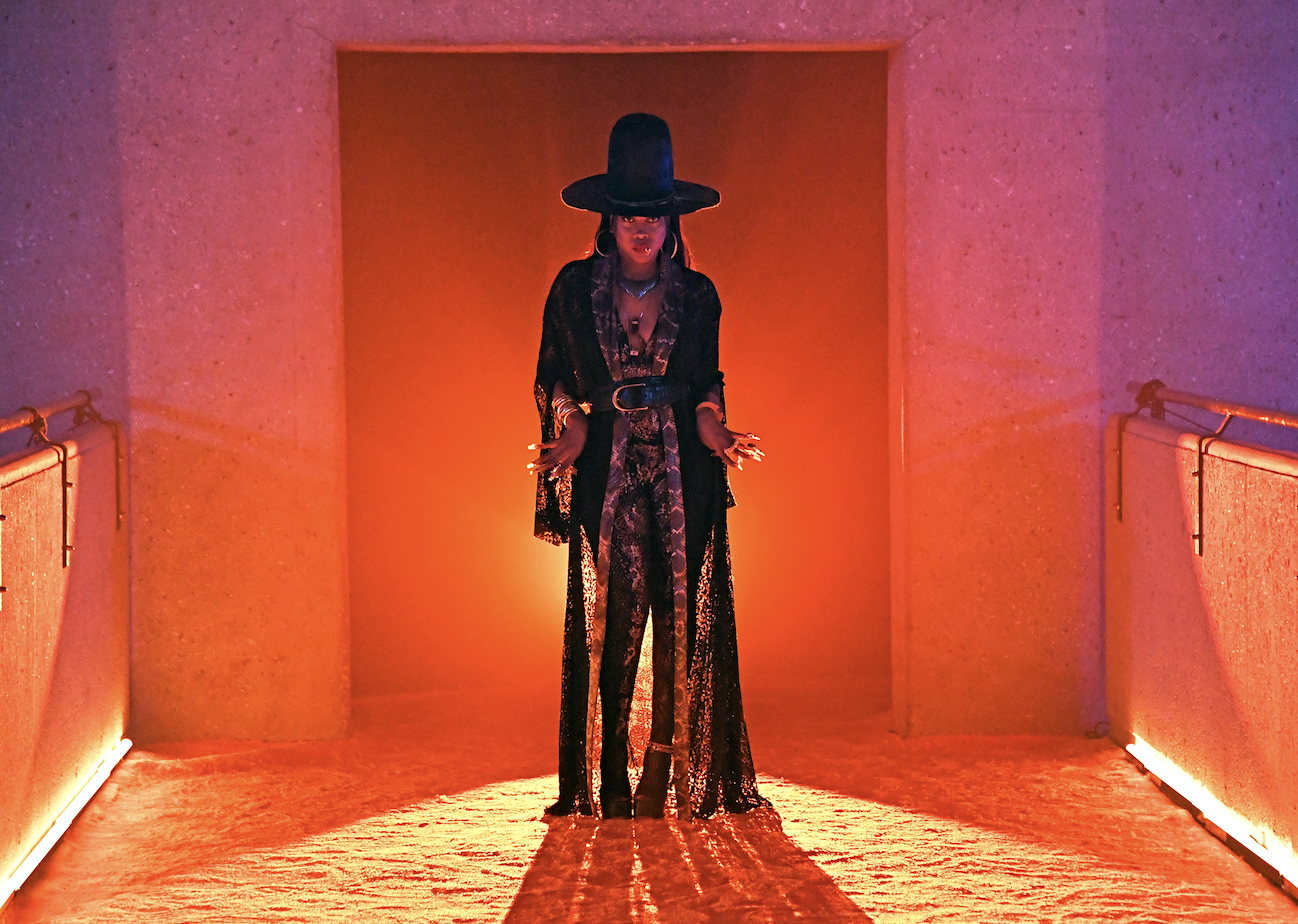Erykah is wearing a floor-length patterned duster jacket with pants, boots, and a hat