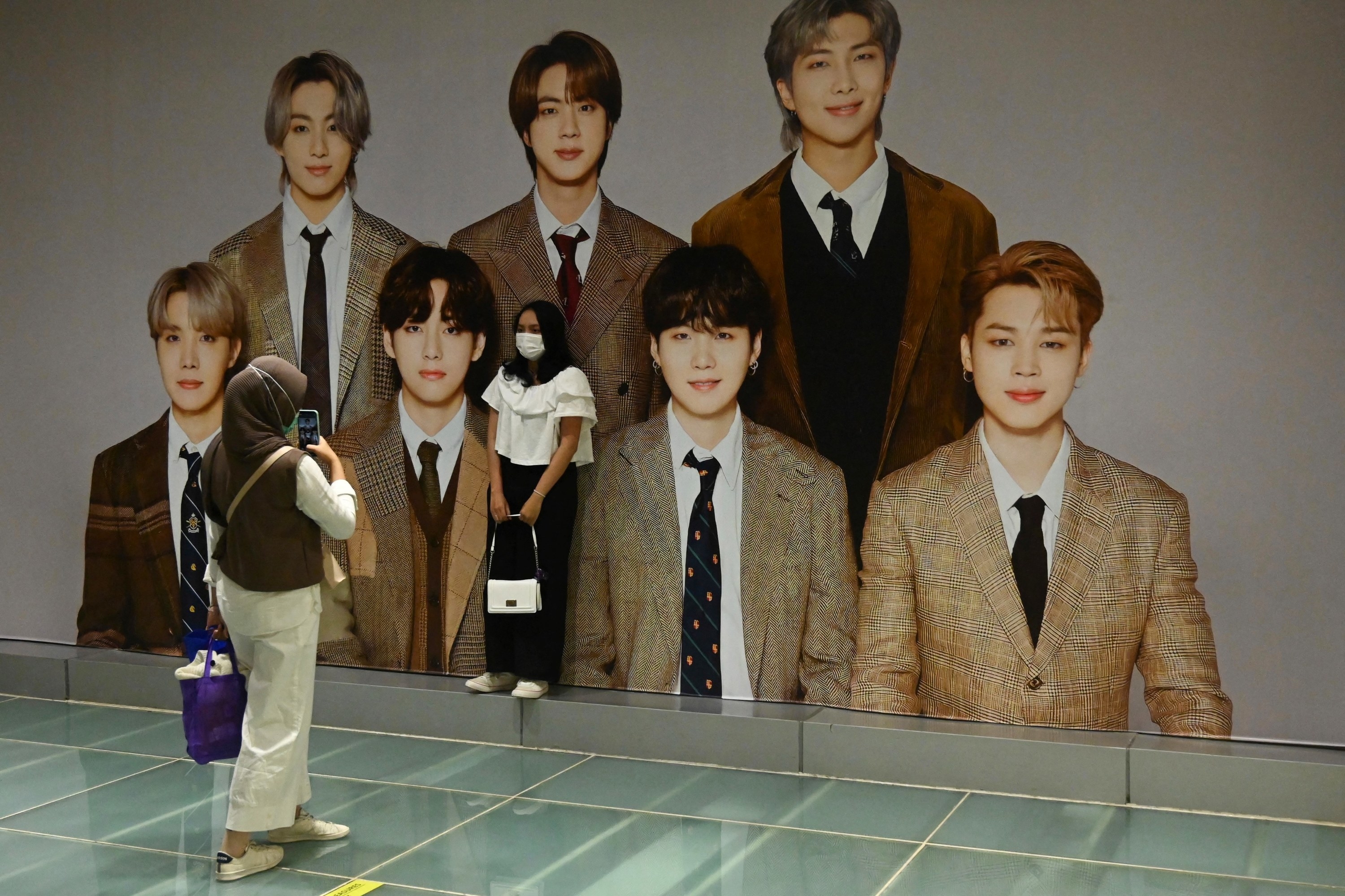 A person taking a photo of someone else as they pose next to a mural of BTS