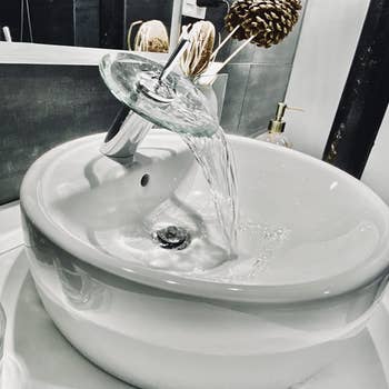 closeup of reviewer's raised bowl bathroom sink. the faucet spouts water from the top that cascades off a clear dish. 