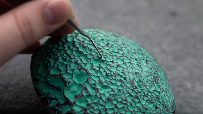 close-up of a textured rock with green paint inside the holes and a picker tool scraping paint out of the holes