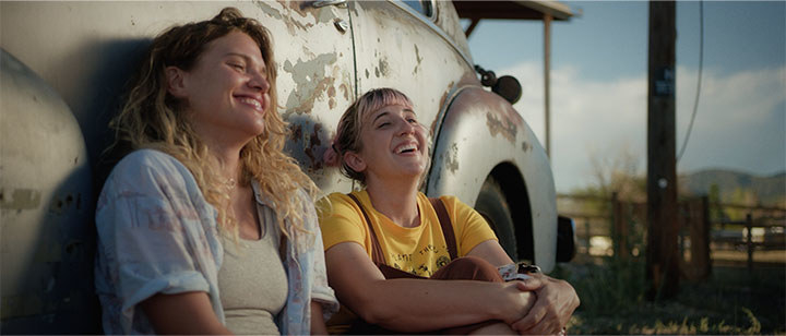 Two women seated against an old car, laughing.