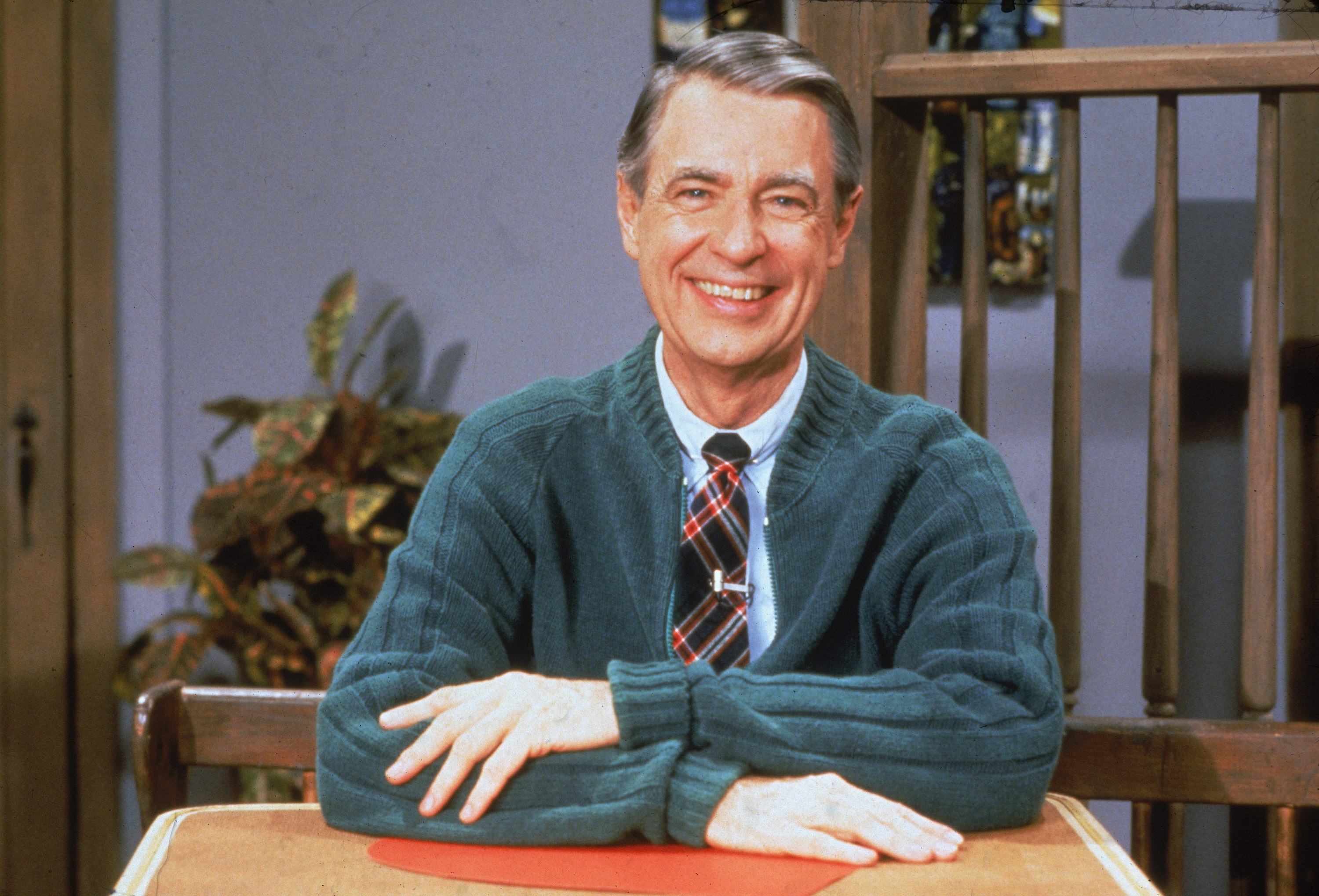 Mr. Rogers sits at a desk with his signature cardigan and dons a bright smile.