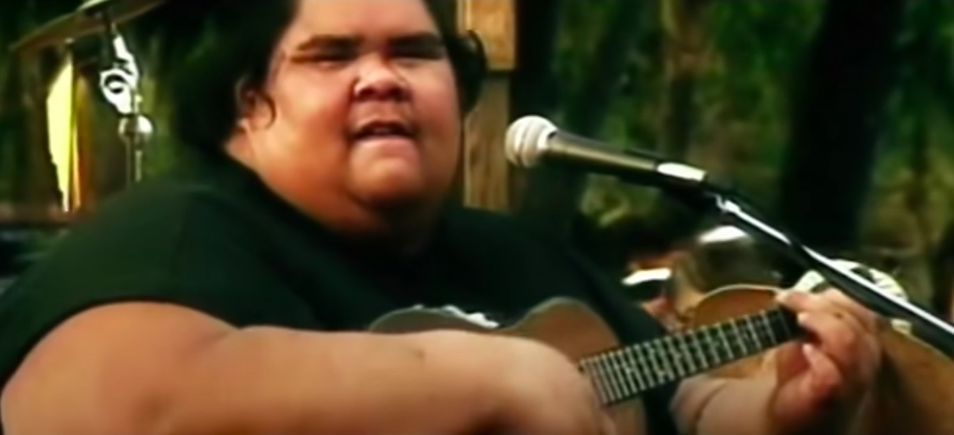 Israel Kamakawiwo&#x27;ole playing a ukelele and singing to a mic.