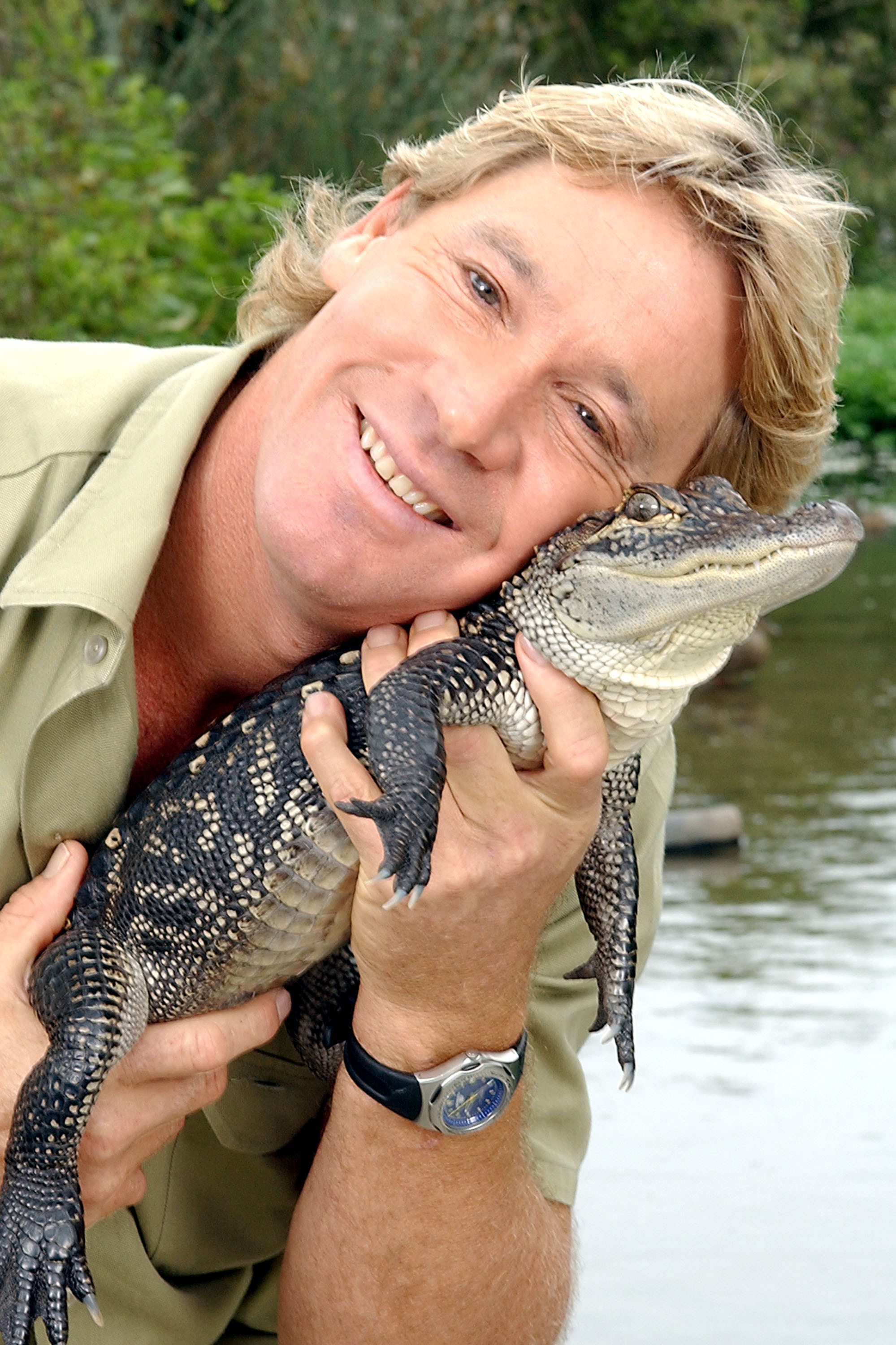 Steve Irwin holds a small alligator up to his face while smiling in front of a body of water.