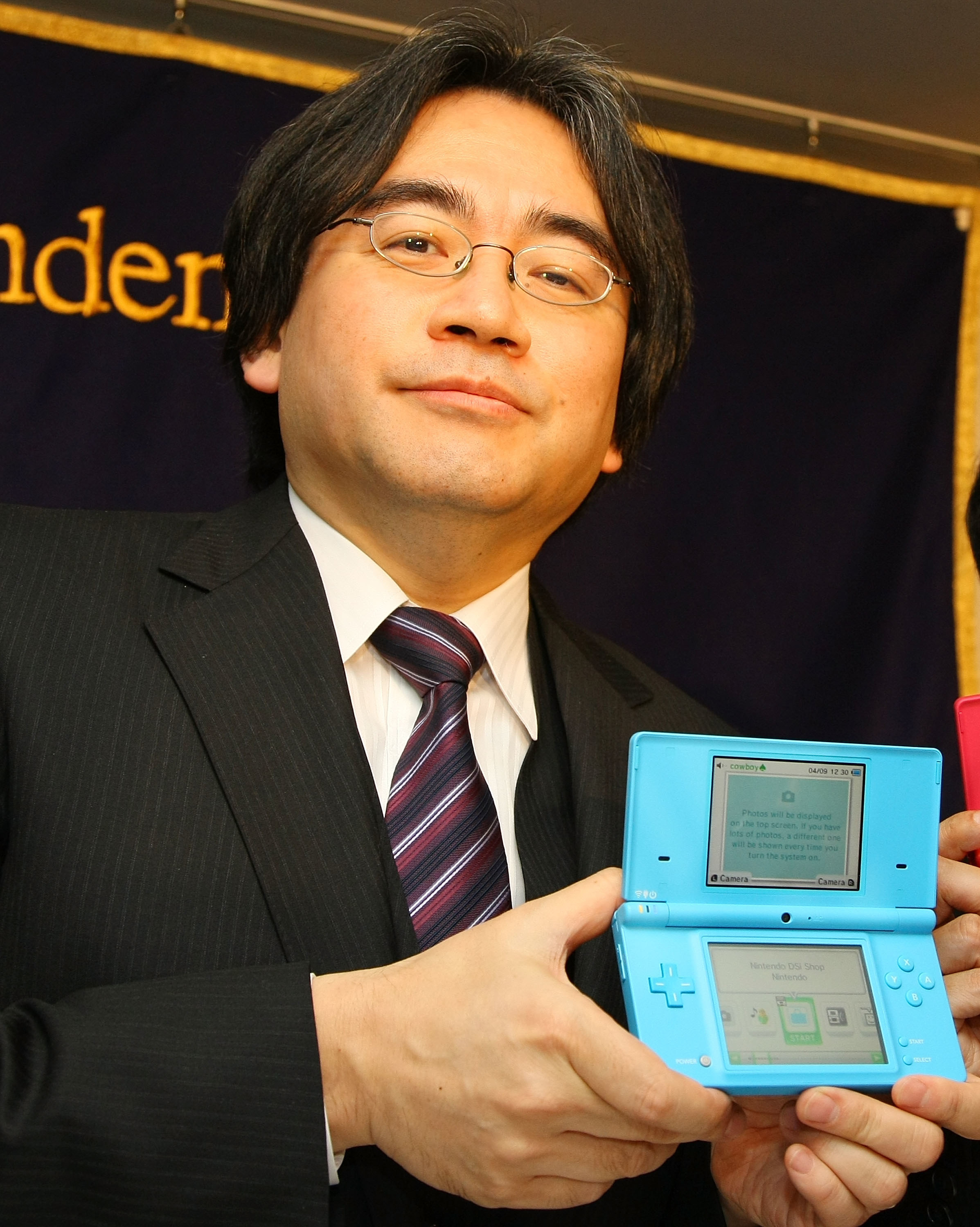 Dressed in a suit, Satoru Iwata proudly holds up a Nintendo DS.