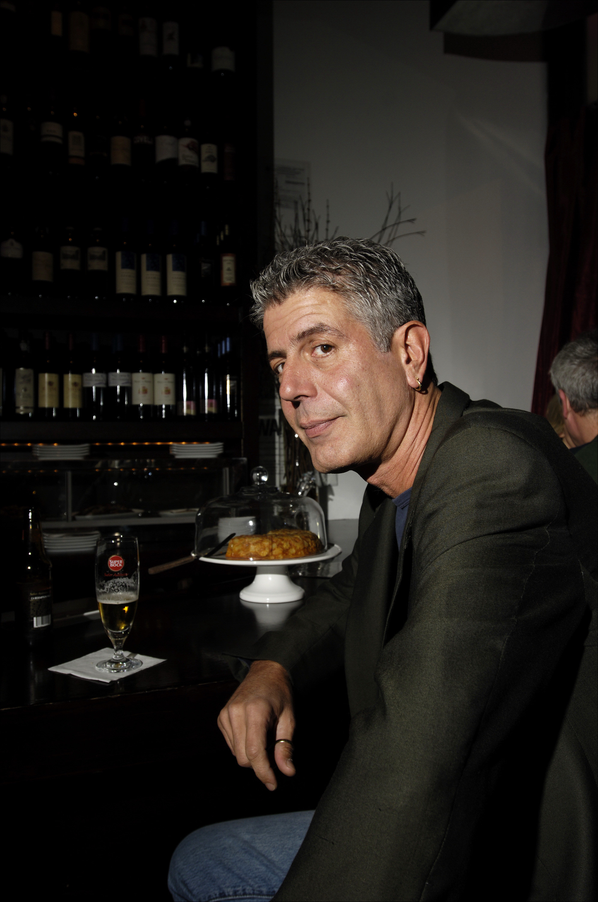 Anthony Bourdain sits at a table with a half-drunk glass of beer and looks to his left at the camera.