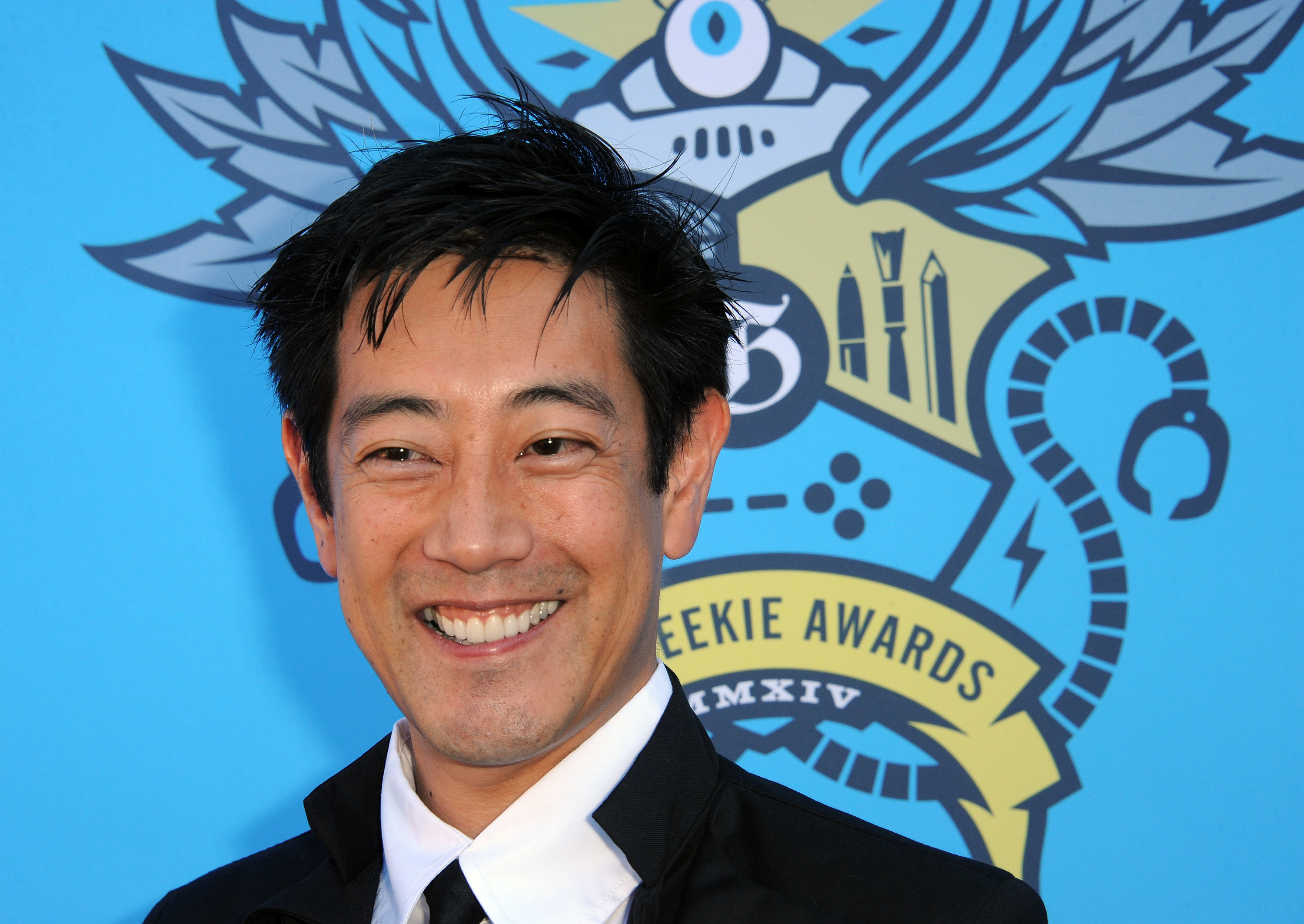 Grant Imahara smiles widely wearing a suit.