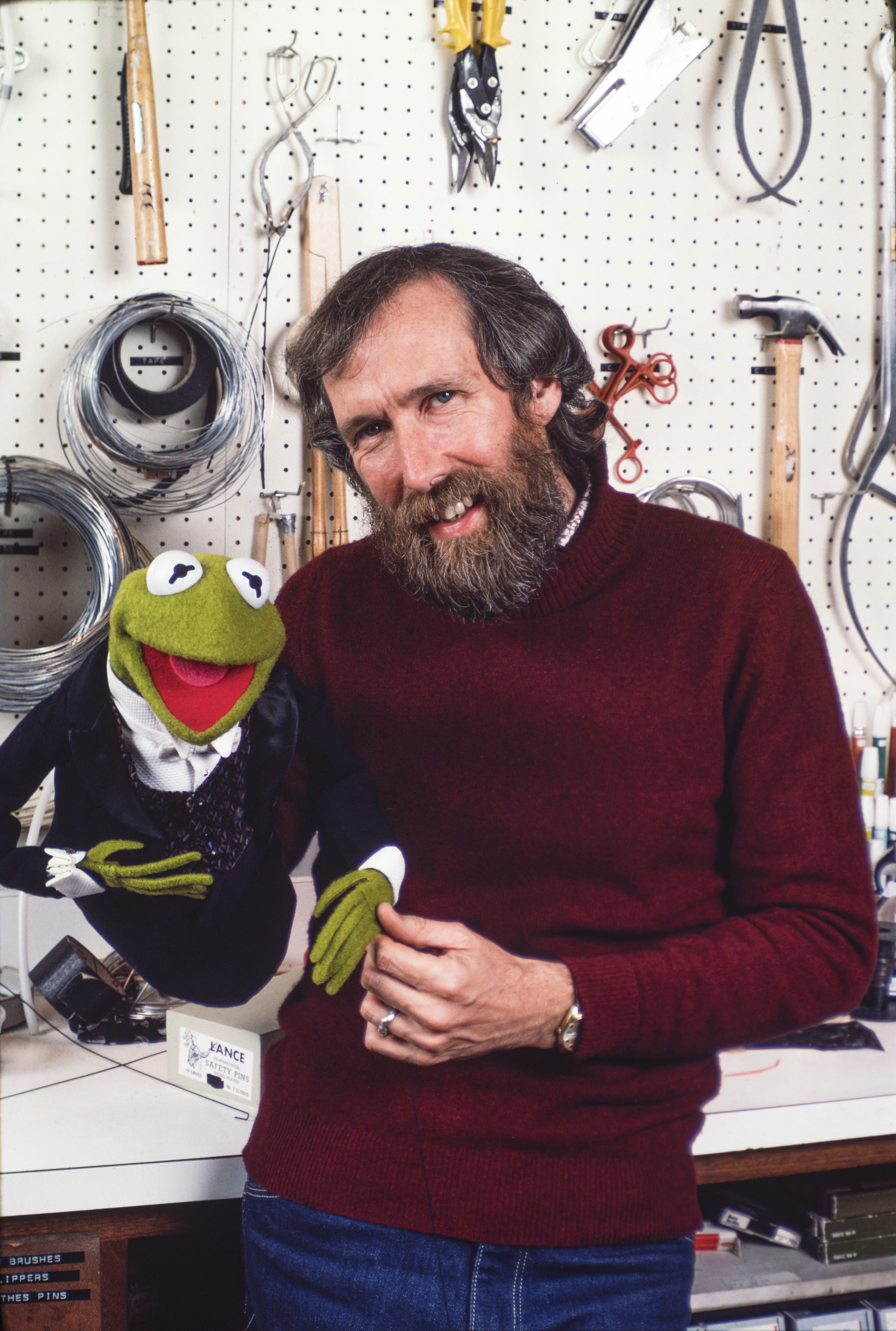 Jim Henson holds a muppet with his right hand while he poses in front of a toolshop.