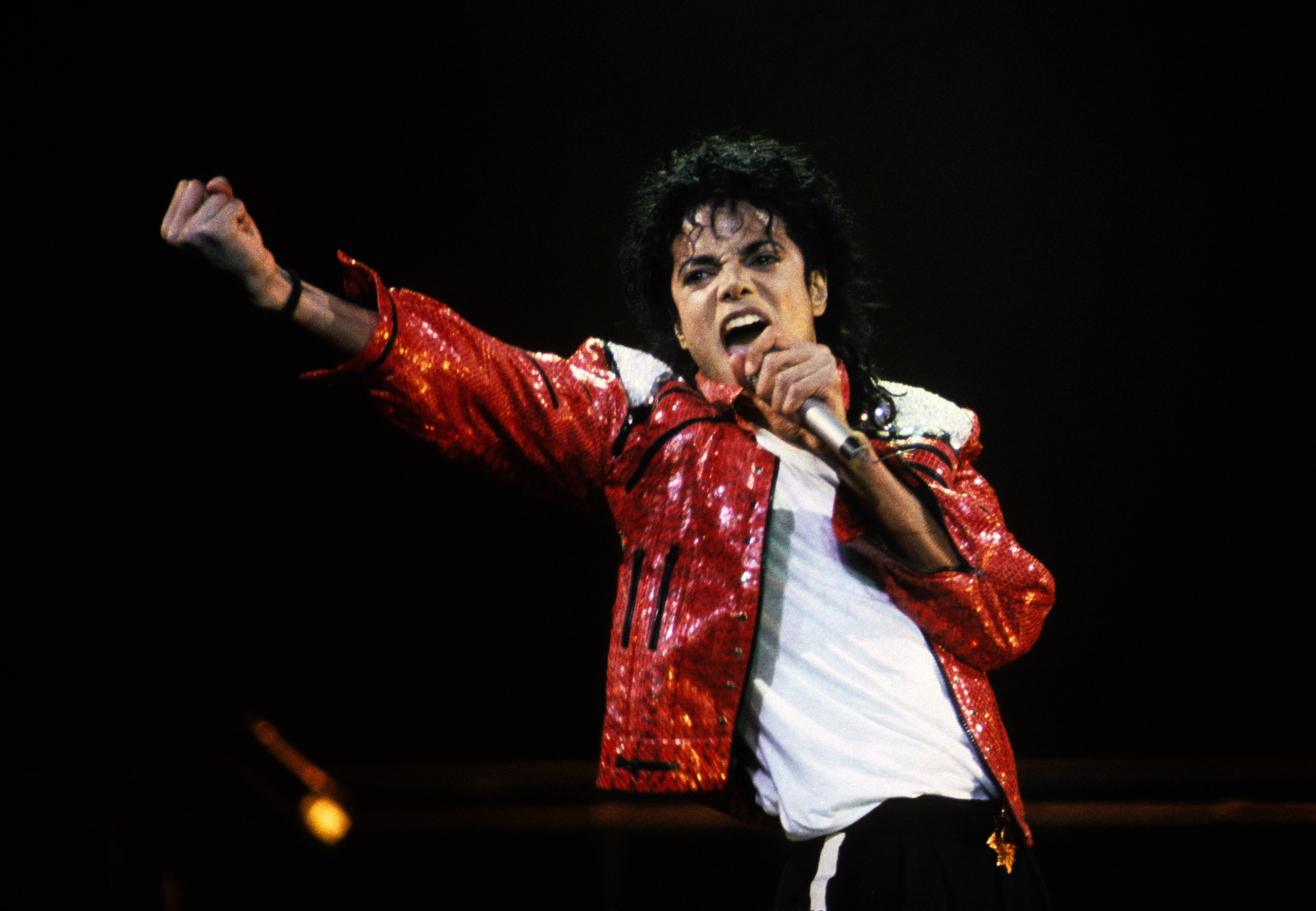 Michael Jackson passionately pumps his fist in the air while his left hand clutches his mic to his mouth.