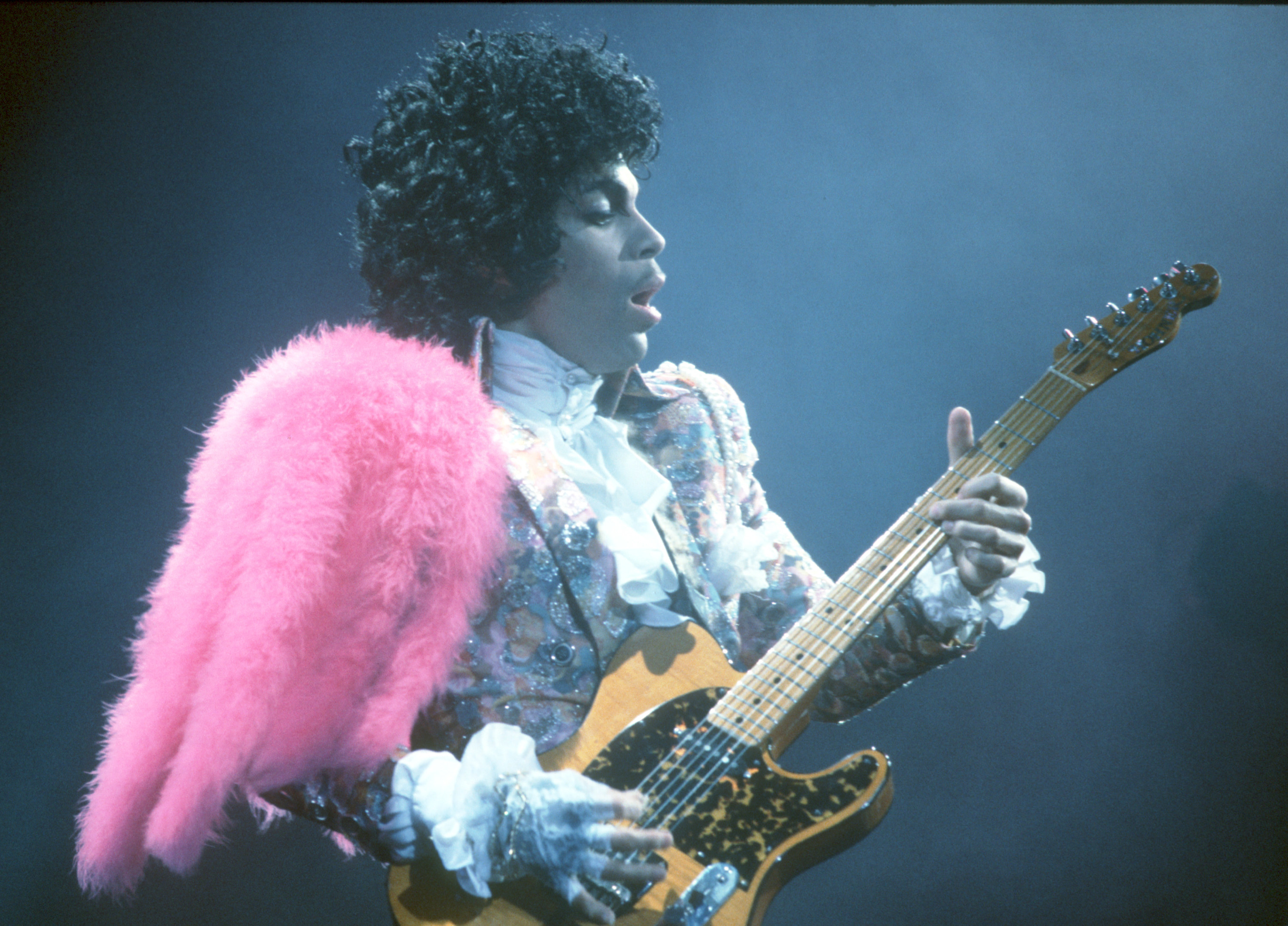 Prince wears a bright pink wing while he plays the electric guitar.