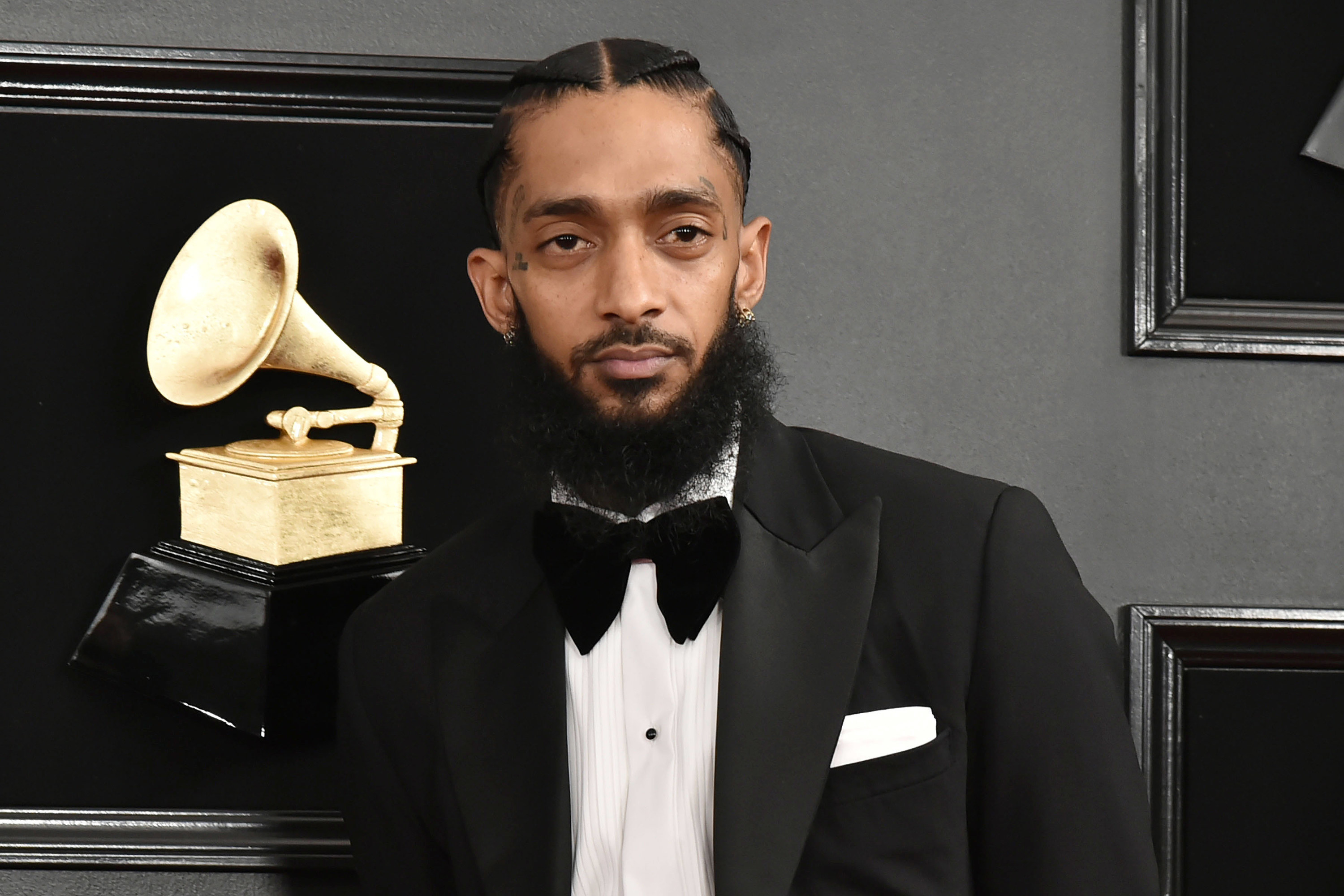 Nipsey Hussle wears a tuxedo and poses elegantly at an event.