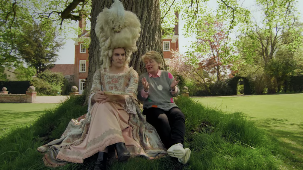 Noel Fielding dressed as Marie Antoinette as he sits against a tree with another person