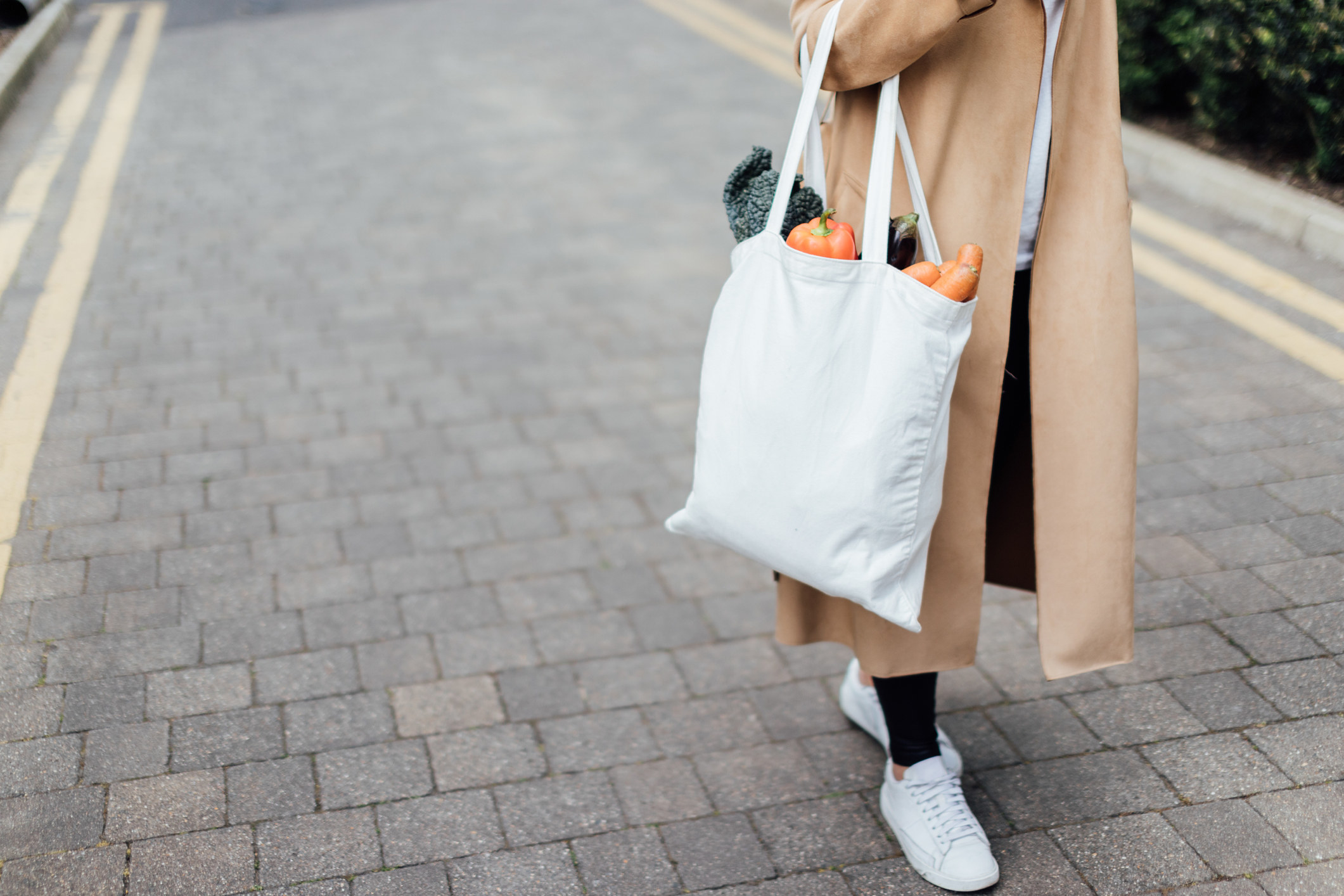 A person wearing a long coat, pants, and sneakers as they carry a reusable bag filled with groceries