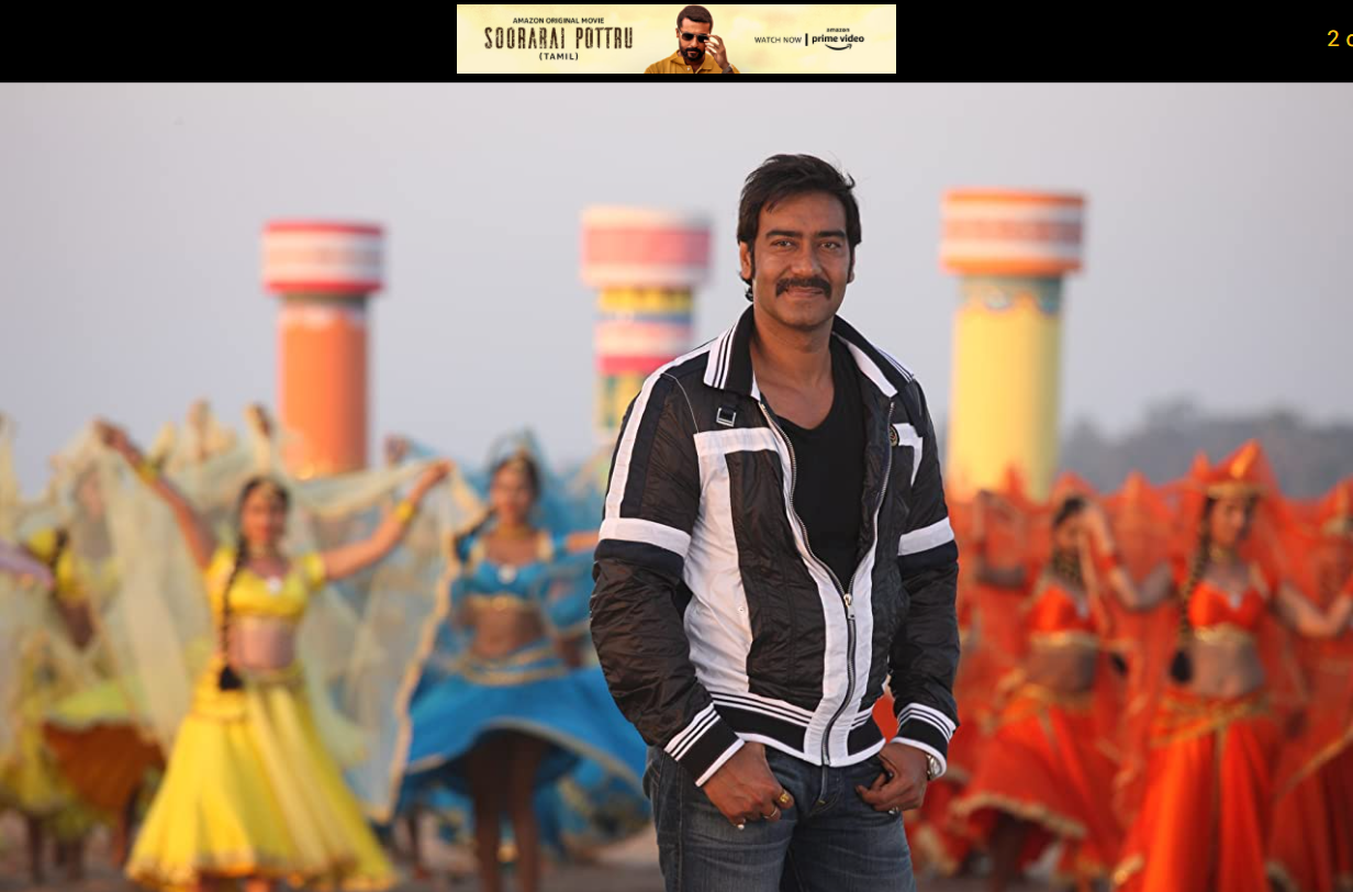 Ajay Devgan standing in front of several women dancing in the background with his thumbs in his pocket