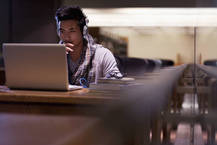 A student sitting at a library desk looking at their laptop with headphones on