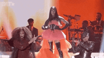 Gif of Mj voguing on stage, she wears a tule pink and orange dress with leather thigh-highs and corset