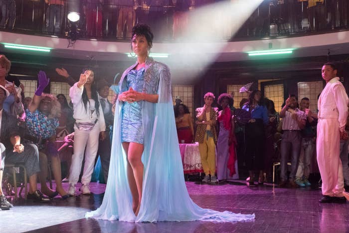 Still from "Pose" featuring Mj at the center in a light blue mini dress and long shawl, there are people behind her in the ballroom watching her