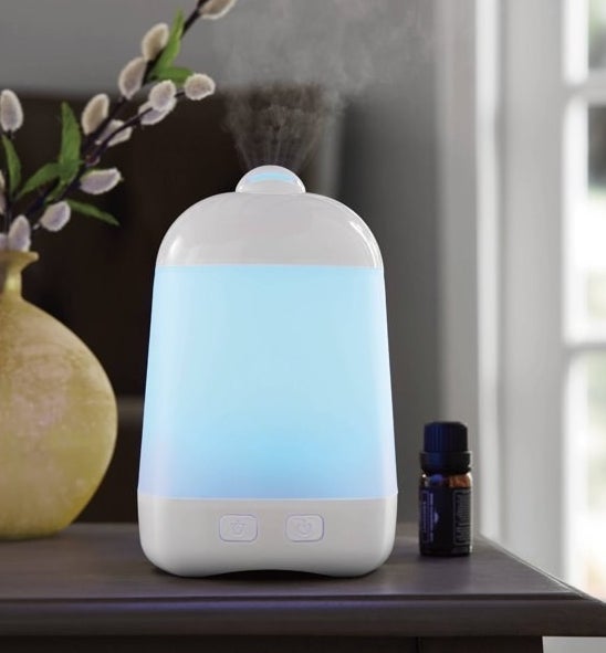 Diffuser with blue light on, sitting on a surface with an essential oil bottle and a flower vase next to it.