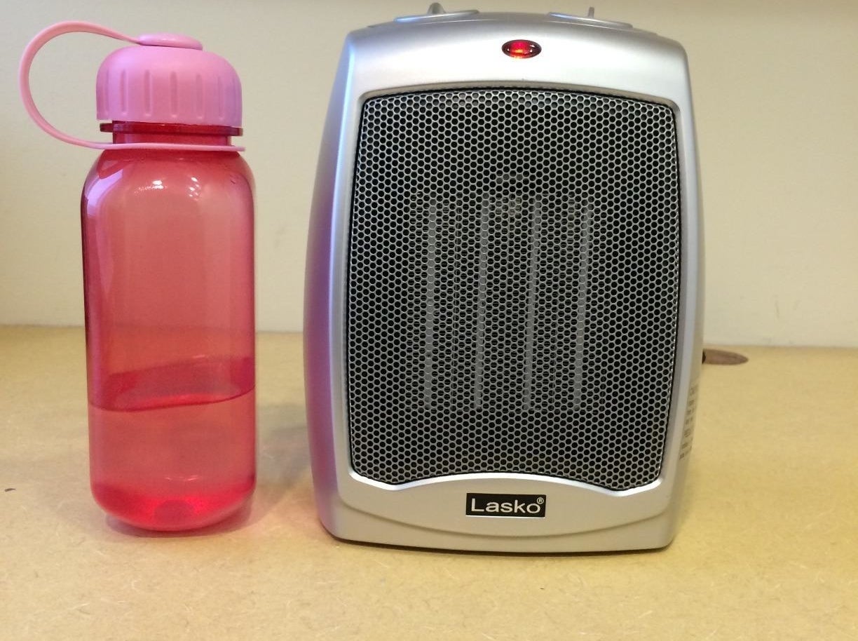the silver space heater next to a pink water bottle to show how small the space heater is