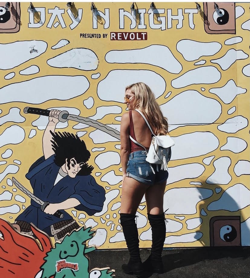 Sam posing in front of a mural wearing denim shorts, boots, and a tank top