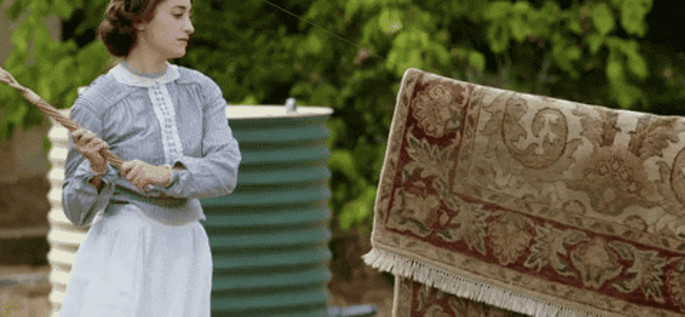 person in old attire dusting a rug by beating it against a wash line