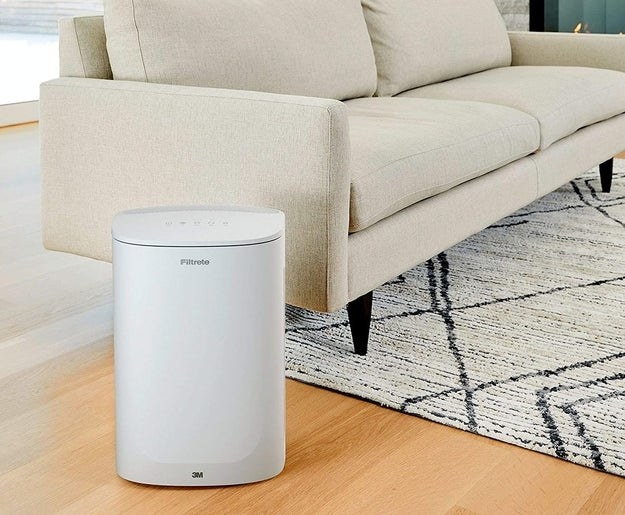 A white air purifier about the size of a of medium trash can next to a couch in a living room