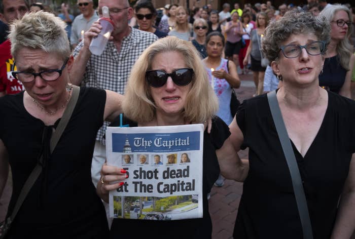 A woman weeping behind sunglasses stands between two other women in a crowd and carries a newspaper that reads &quot;5 shot dead at the Capital&quot;