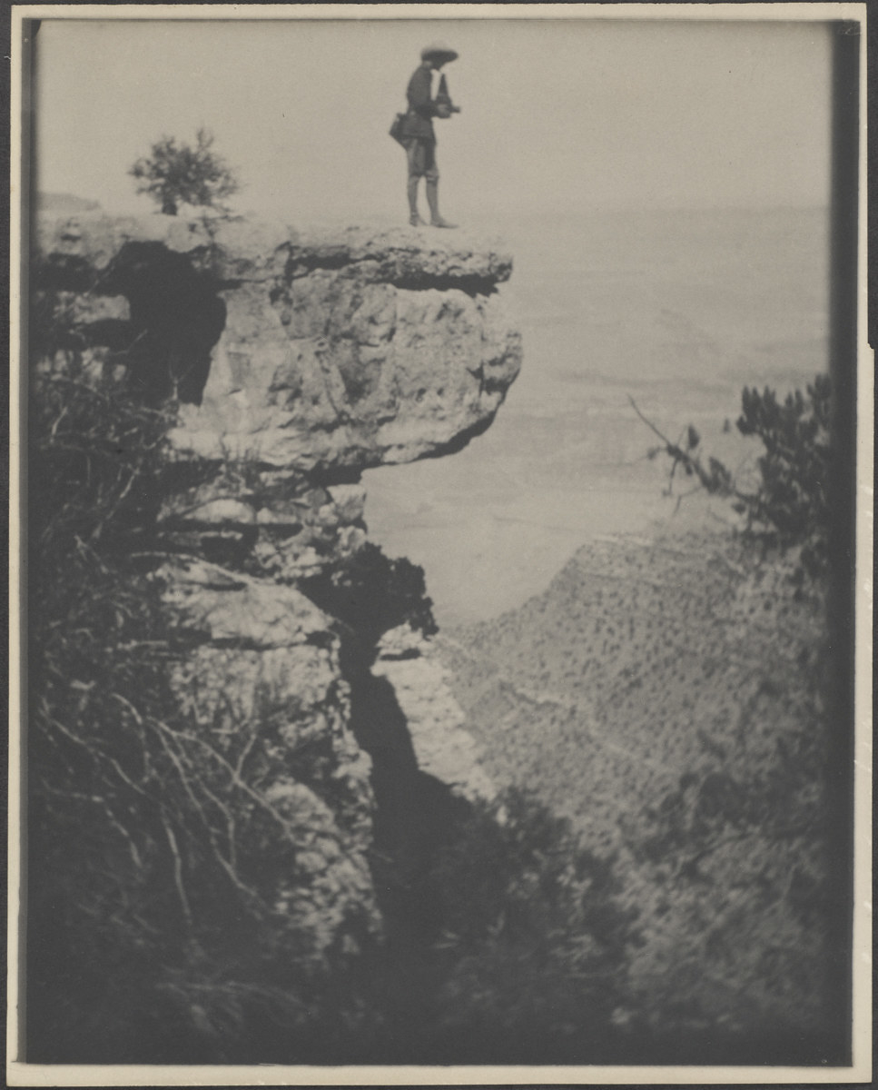 A person in a hat stands on a steep ledge overlooking the Grand Canyon, holding a camera