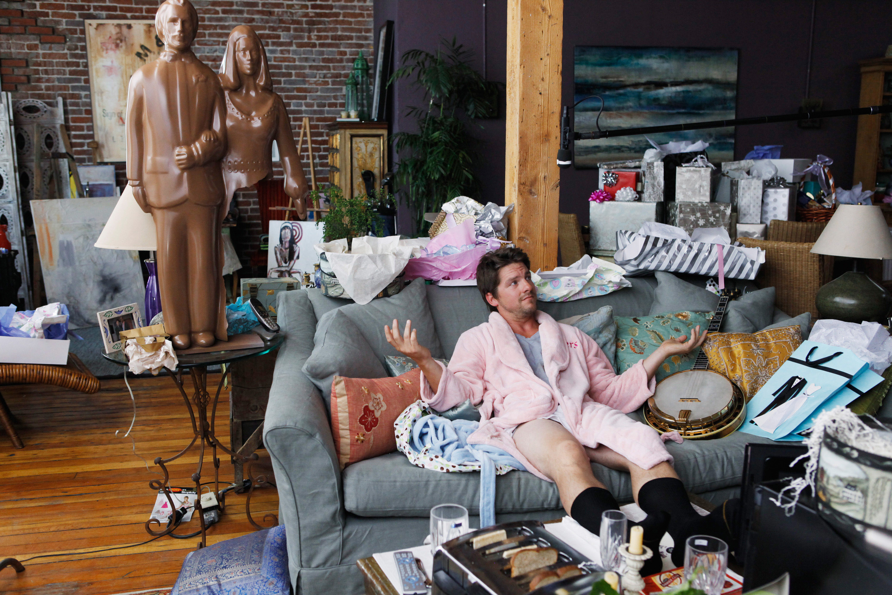 Zachary Knighton shrugging in a pink bathrobe while sitting on the couch.