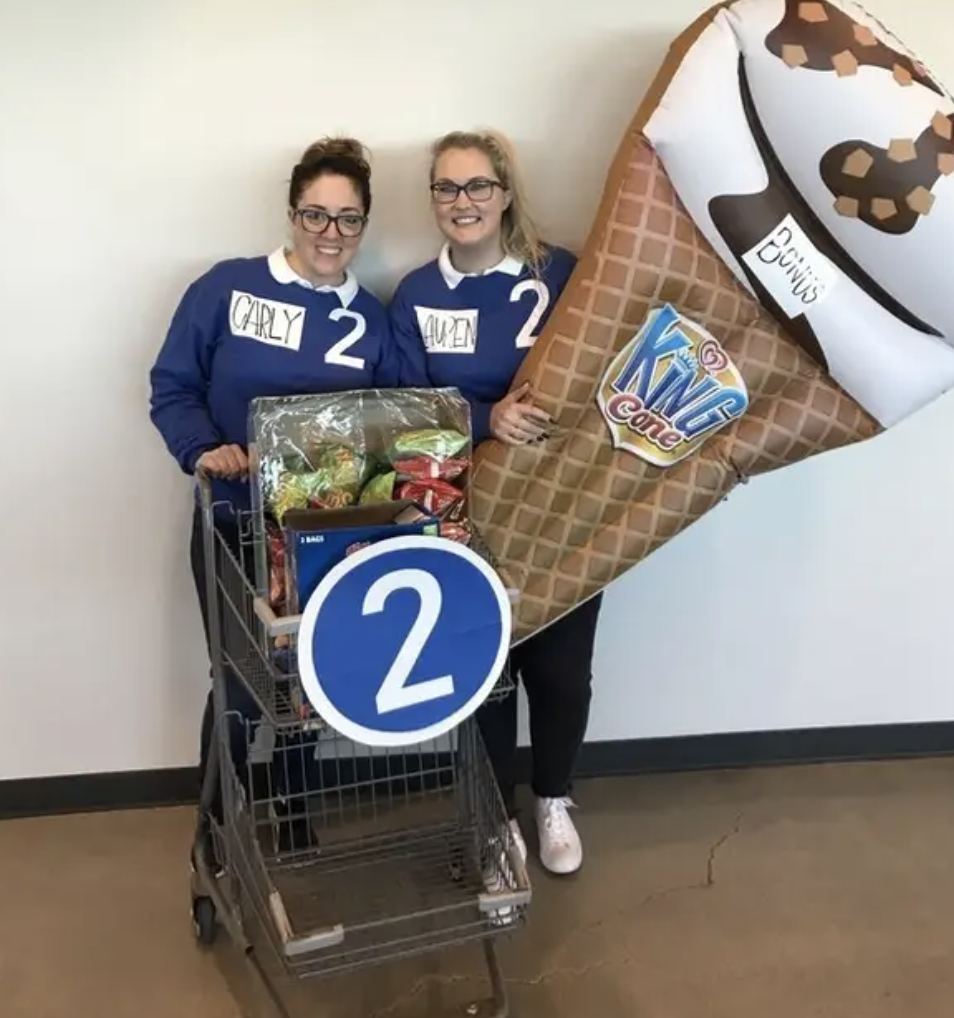 Two people wearing matching sweaters with name tags while pushing a shopping cart and carrying a giant ice cream inflatable