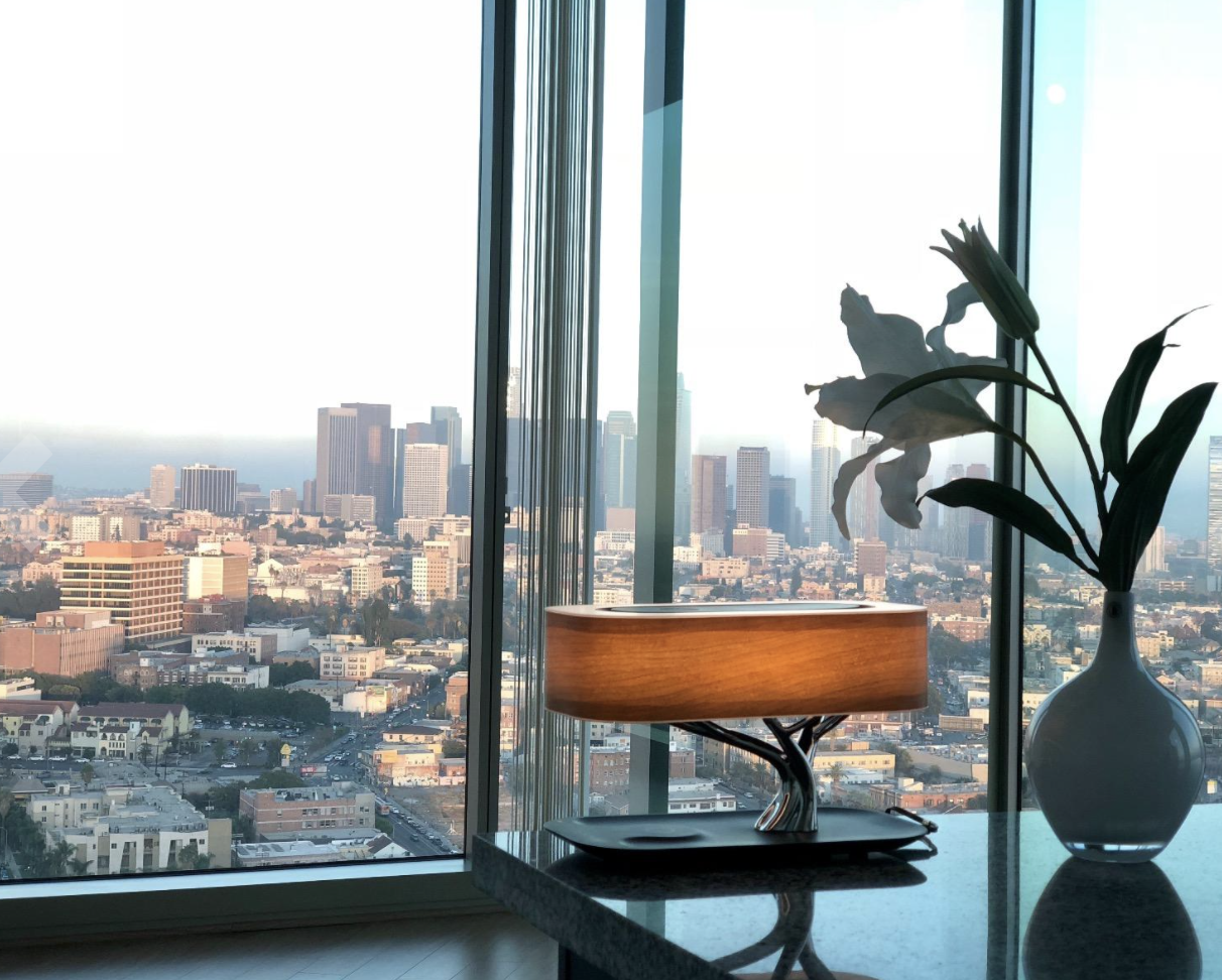 lamp sitting on a desk in an office overlooking a big city. The lamp has an oval wooden shade that looks like the top portion of a tree, a lamp stem that looks like the tree trunk, and a base where the phone can lay and charge wirelessly