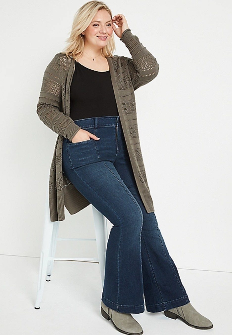 model wearing the flare jeans sitting on a white chair with a black top and green cardigan
