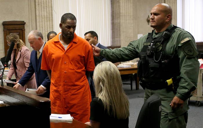 R. Kelly in an orange prison uniform and his hands behind him in a courtroom