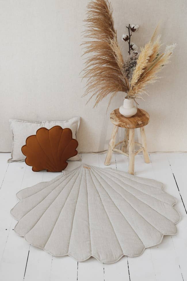 sea shell shaped mat on wooden floor. it's beside matching pillows and dried flowers.