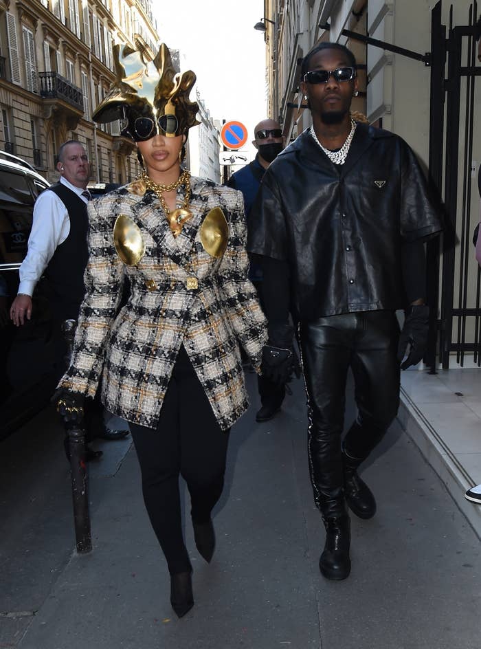 Cardi and Offset walking hand in hand on the street, with Cardi wearing a plaid jacket with gold accents and a gold headpiece, and Offset in black leather