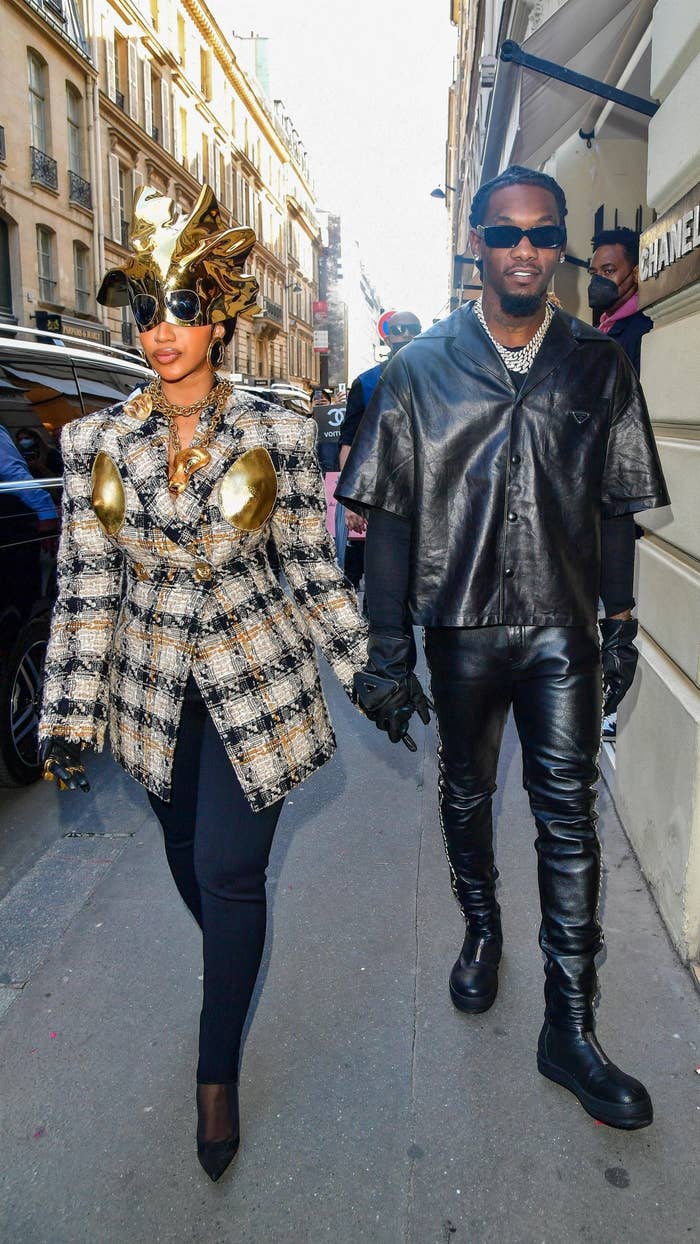 Cardi and Offset walking hand in hand on the street, with Cardi wearing a plaid jacket with gold accents and a gold headpiece, and Offset in black leather