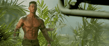 A shirtless man stands in the middle of a forest, while the wind blows around him