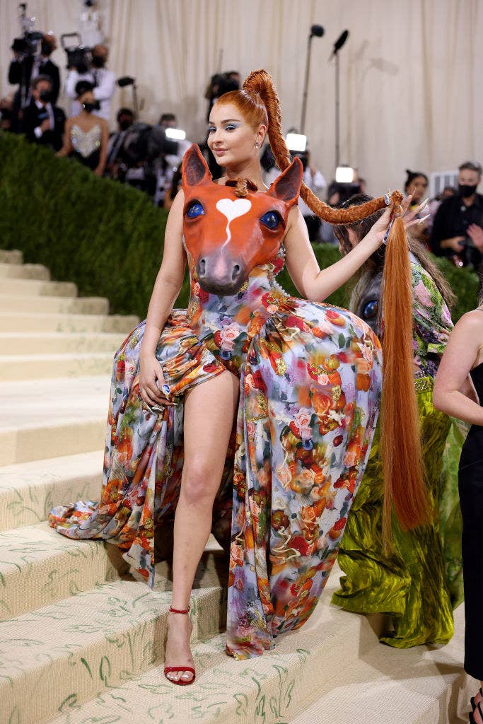 Kim Petras wearing a dress with a floral skirt and a horse head on the bodice while she clutches a floor-length ponytail in one hand