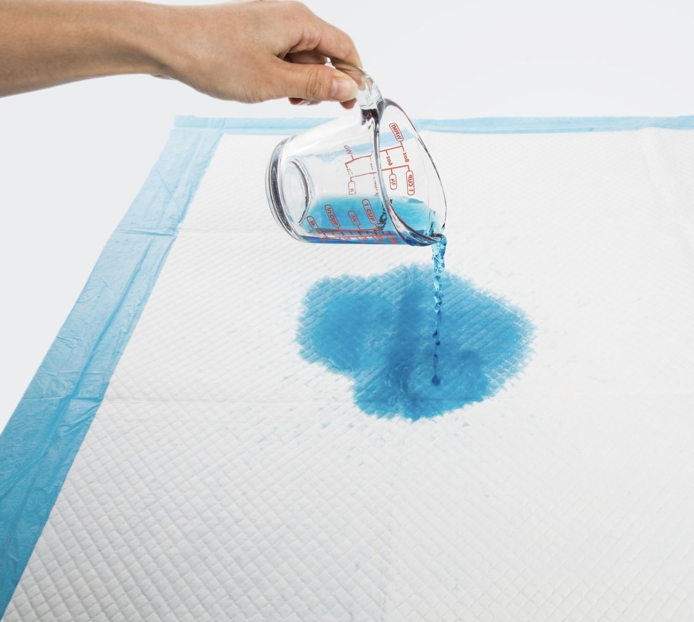 The pad is white with a blue border and glass measuring cup is pouring blue liquid onto the fabric