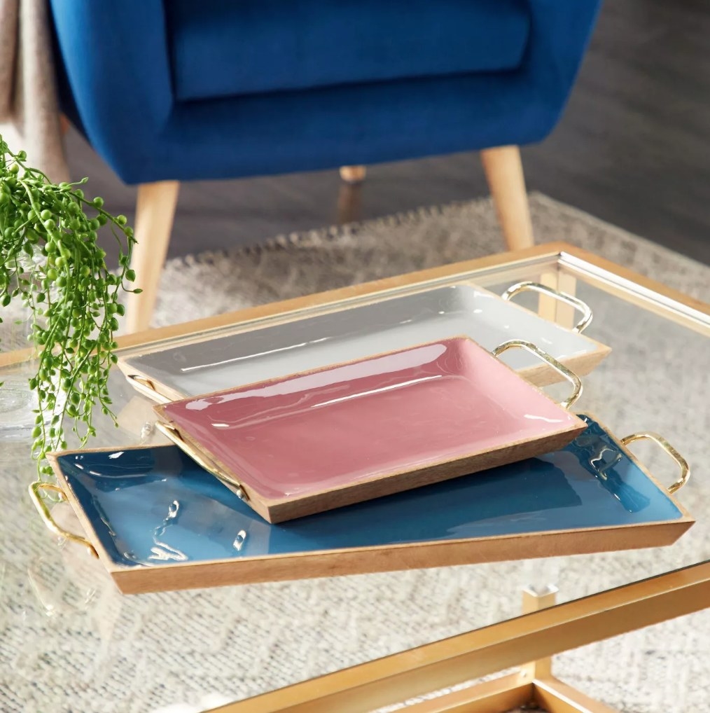 A set of 3 (pink/grey/blue), wood enamel serving trays with gold handles on a coffee table