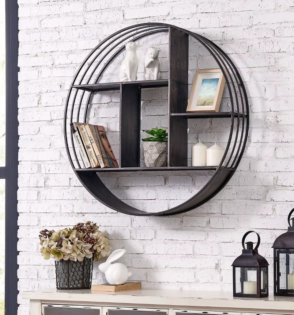 A circular, metallic grey, industrial wall shelf filled with books, frames, candles, and other decor
