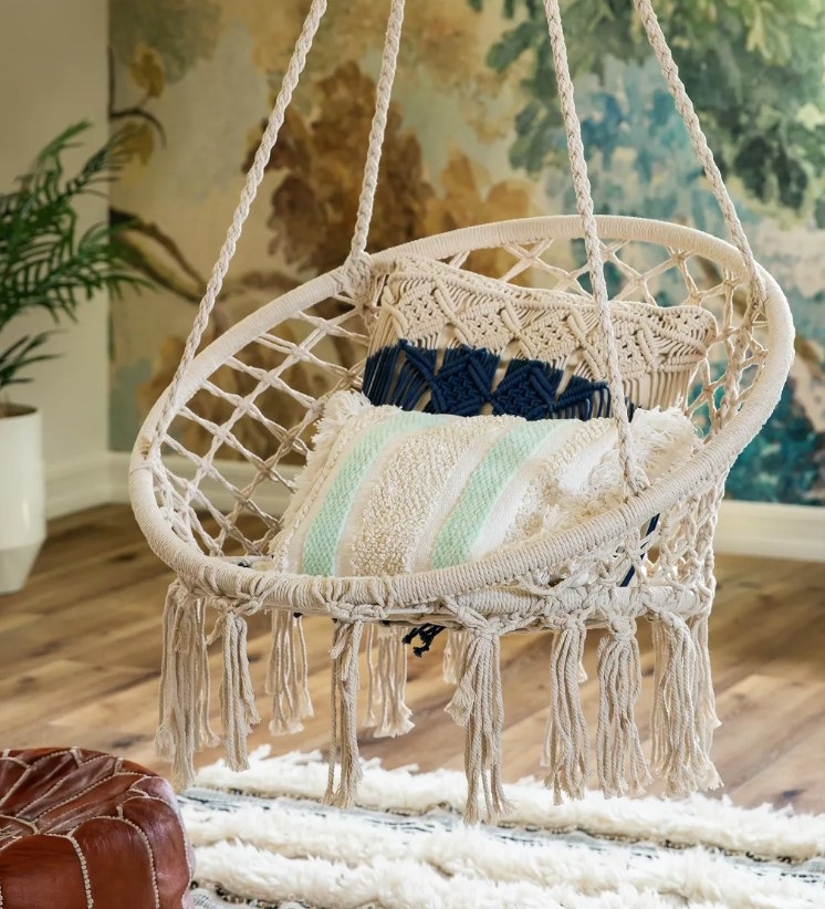 A beige, hanging, macrame swing chair in a living room with throw pillows atop
