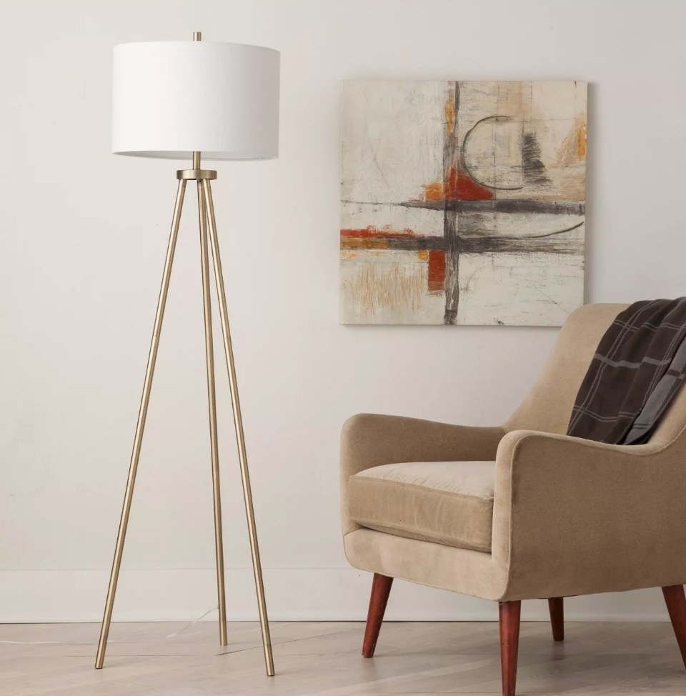 A brass, tripod floor lamp with a white shade next to an accent chair